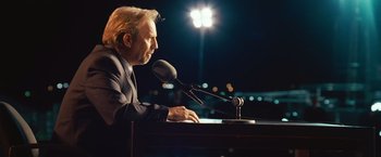Movie still from “Swing Vote” (2008), directed by Joshua Michael Stern – A man sitting in front of a microphone at a podium; Close Up shot, Over the shoulder angle