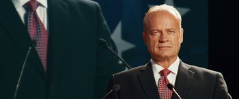 Movie still from “Swing Vote” (2008), directed by Joshua Michael Stern – A man in a black suit and a red tie standing in front of a microphone; Medium shot, Low angle