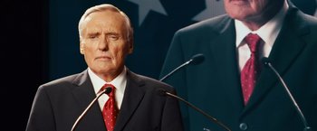 Movie still from “Swing Vote” (2008), directed by Joshua Michael Stern – An older man wearing a suit and tie while standing at a podium with microphones; Close Up shot, Low angle