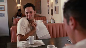 Movie still from “Swingers” (1996), directed by Doug Liman – A man sitting at a table holding a paper bag; Medium shot, Over the shoulder angle