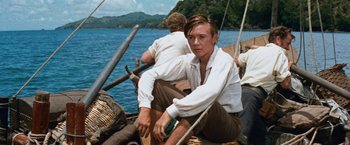 Movie still from “Swiss Family Robinson” (1960), directed by Ken Annakin – A man sitting on the side of a boat in the water; Medium shot, Low angle