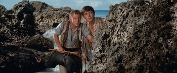 Movie still from “Swiss Family Robinson” (1960), directed by Ken Annakin – Two young men standing next to each other on a beach; Medium shot, Low angle