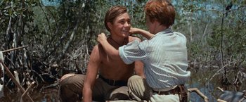 Movie still from “Swiss Family Robinson” (1960), directed by Ken Annakin – A man fixing another man's collar while sitting on the ground; Medium shot, Over the shoulder angle