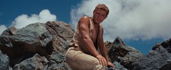 Movie still from “Swiss Family Robinson” (1960), directed by Ken Annakin – A man sitting on top of a rock on top of a mountain; Medium shot, Low angle