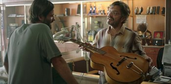 Movie still from “Sword of Trust” (2019), directed by Lynn Shelton – A man holding a guitar in a room with a man standing next to him; Medium shot, Over the shoulder angle