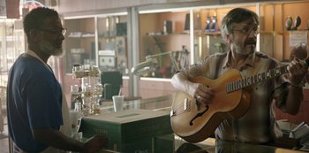 Movie still from “Sword of Trust” (2019), directed by Lynn Shelton – A man sitting at a table playing a guitar; Medium shot, Over the shoulder angle