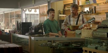 Movie still from “Sword of Trust” (2019), directed by Lynn Shelton – A man standing in front of a counter next to another man; Medium shot, Over the shoulder angle