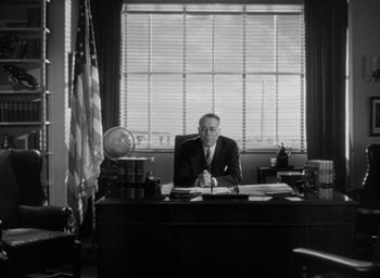 Movie still from “T-Men” (1947), directed by Anthony Mann – A man sitting at a desk in front of an american flag; Wide shot, Low angle