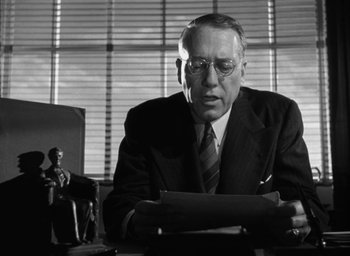 Movie still from “T-Men” (1947), directed by Anthony Mann – An older man in a suit and tie sitting at a desk; Medium shot, Low angle