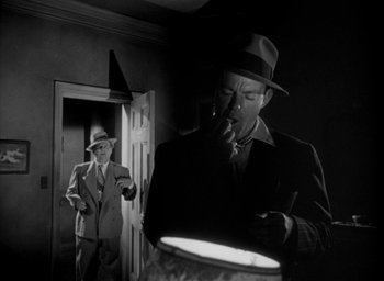 Movie still from “T-Men” (1947), directed by Anthony Mann – A man smoking a cigarette while another man is standing next to a lamp; Medium shot, Low angle