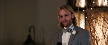 Movie still from “Table 19” (2017), directed by Jeffrey Blitz – A man with long hair and a bow tie is standing in front of a microphone; Close Up shot, Over the shoulder angle