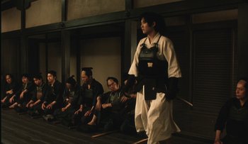Movie still from “Taboo” (1999), directed by Nagisa Ôshima – A group of people sitting in a row watching a performer; Wide shot, Low angle