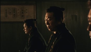 Movie still from “Taboo” (1999), directed by Nagisa Ôshima – Two asian men standing next to each other in a dark room; Medium shot, Over the shoulder angle