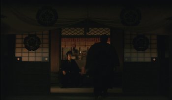 Movie still from “Taboo” (1999), directed by Nagisa Ôshima – A man sitting on the ground in front of a building at night; Wide shot, Over the shoulder angle