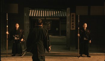 Movie still from “Taboo” (1999), directed by Nagisa Ôshima – A man in a black jacket is standing in front of a building; Wide shot, Over the shoulder angle
