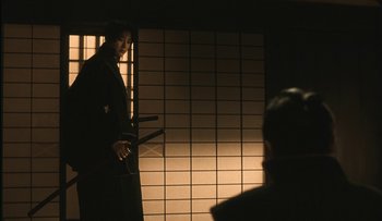 Movie still from “Taboo” (1999), directed by Nagisa Ôshima – A man standing in front of a wall holding a pair of katanas; Medium shot, Over the shoulder angle