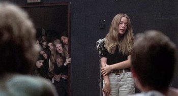 Movie still from “Taking Off” (1971), directed by Milos Forman – A woman standing at a microphone in front of a group of people; Medium shot, Over the shoulder angle
