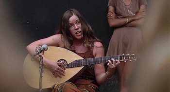 Movie still from “Taking Off” (1971), directed by Milos Forman – A woman holding a guitar while standing next to a microphone; Medium shot, Low angle