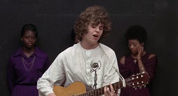 Movie still from “Taking Off” (1971), directed by Milos Forman – A young man is playing an acoustic guitar in front of a microphone; Medium shot, Low angle