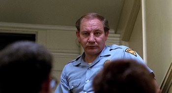 Movie still from “Taking Off” (1971), directed by Milos Forman – A man in a police uniform standing in front of a group of people; Close Up shot, Low angle