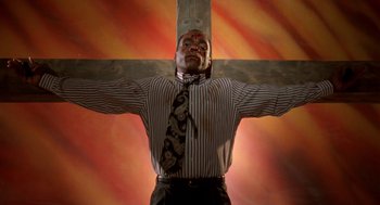 Movie still from “Tales from the Hood” (1995), directed by Rusty Cundieff – A man in a striped shirt and a neck tie with his arms outstretched in front of a wooden cross; Medium shot, Low angle