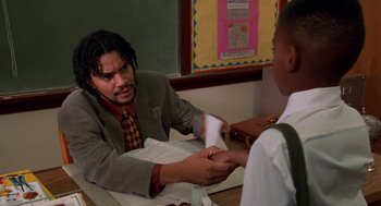 Movie still from “Tales from the Hood” (1995), directed by Rusty Cundieff – A man sitting at a table with another man holding a piece of paper in front of him; Medium shot, High angle
