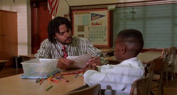 Movie still from “Tales from the Hood” (1995), directed by Rusty Cundieff – Two people sitting at a table in front of a blackboard; Medium shot, Low angle