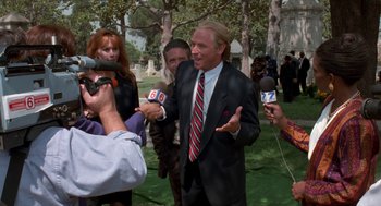 Movie still from “Tales from the Hood” (1995), directed by Rusty Cundieff – A man in a black suit is being interviewed by reporters; Medium shot, Over the shoulder angle