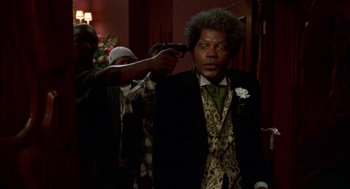 Movie still from “Tales from the Hood” (1995), directed by Rusty Cundieff – A man in a suit and tie holding a gun; Medium shot, Over the shoulder angle