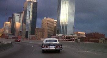 Movie still from “Talk Radio” (1988), directed by Oliver Stone – A car driving down a street near tall buildings; Extreme Wide shot, High angle