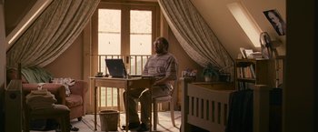 Movie still from “Tamara Drewe” (2010), directed by Stephen Frears – A man sitting in front of a laptop computer on top of a desk; Wide shot, Low angle