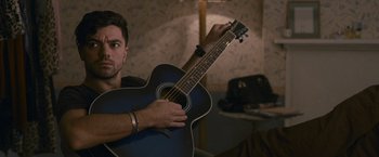 Movie still from “Tamara Drewe” (2010), directed by Stephen Frears – A man holding a blue guitar in a room; Medium shot, Over the shoulder angle