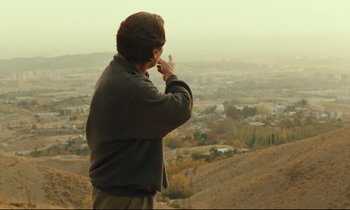 Movie still from “Taste of Cherry” (1997), directed by Abbas Kiarostami – A man standing on top of a hill looking at a city; Medium shot, Over the shoulder angle