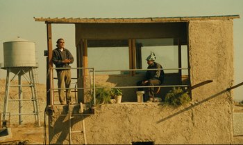 Movie still from “Taste of Cherry” (1997), directed by Abbas Kiarostami – Two men are standing on a balcony with potted plants; Wide shot, Low angle