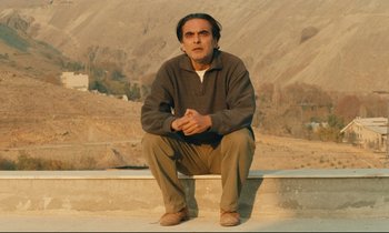 Movie still from “Taste of Cherry” (1997), directed by Abbas Kiarostami – A man squatting on a ledge in front of mountains; Medium shot, Low angle