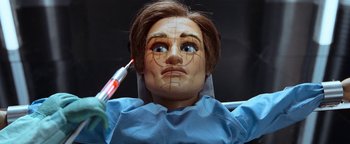 Movie still from “Team America: World Police” (2004), directed by Trey Parker – A doll is being held up with a syringe in it's mouth; Extreme Close Up shot, Overhead angle