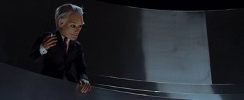 Movie still from “Team America: World Police” (2004), directed by Trey Parker – A man in a black suit is sitting in front of a metal wall; Close Up shot, Low angle