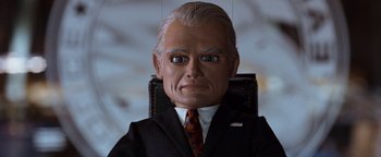 Movie still from “Team America: World Police” (2004), directed by Trey Parker – A man in a suit and tie is sitting in a chair; Close Up shot, Low angle