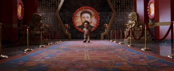 Movie still from “Team America: World Police” (2004), directed by Trey Parker – A small doll standing in front of a large picture of a man; Wide shot, High angle