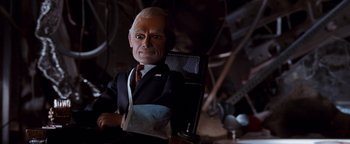 Movie still from “Team America: World Police” (2004), directed by Trey Parker – A man in a suit and tie sitting in a chair; Close Up shot, Low angle