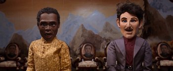 Movie still from “Team America: World Police” (2004), directed by Trey Parker – Two men are standing in front of a painting; Close Up shot, Low angle