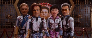 Movie still from “Team America: World Police” (2004), directed by Trey Parker – A group of people standing next to each other in front of a poster; Close Up shot, Low angle