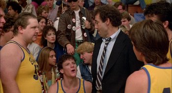 Movie still from “Teen Wolf” (1985), directed by Rod Daniel – A man in a suit and tie standing in front of a group of people; Medium shot, Over the shoulder angle