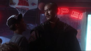 Movie still from “Teenage Mutant Ninja Turtles” (1990), directed by Steve Barron – A man in a black shirt is standing next to another man in a black hat; Medium shot, Low angle