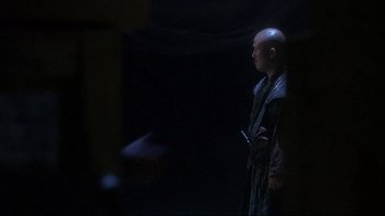 Movie still from “Teenage Mutant Ninja Turtles” (1990), directed by Steve Barron – A bald man in a robe holding a knife in a dark room; Medium shot, Over the shoulder angle