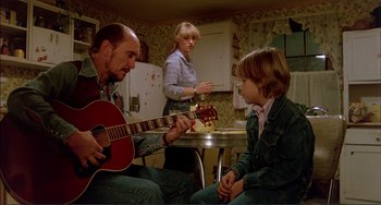 Movie still from “Tender Mercies” (1983), directed by Bruce Beresford – A man playing a guitar in front of a woman and a child; Medium shot, Over the shoulder angle