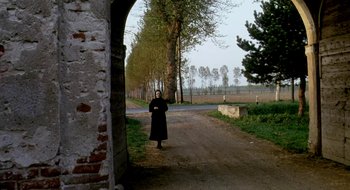 Movie still from “Teorema” (1968), directed by Pier Paolo Pasolini – A woman in a long black coat standing on the side of a road; Wide shot, High angle