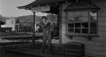 Movie still from “Terror in a Texas Town” (1958), directed by Joseph H. Lewis – A man standing in front of a train station holding a box; Wide shot, Low angle