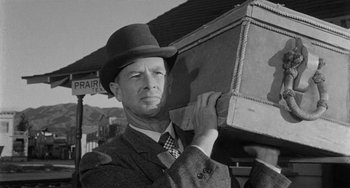 Movie still from “Terror in a Texas Town” (1958), directed by Joseph H. Lewis – A black and white photo of a man in a suit and hat holding a suitcase; Medium shot, Low angle