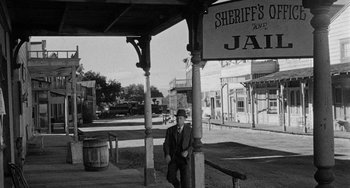 Movie still from “Terror in a Texas Town” (1958), directed by Joseph H. Lewis – A man in a hat standing under an awning in an old western town; Wide shot, Low angle